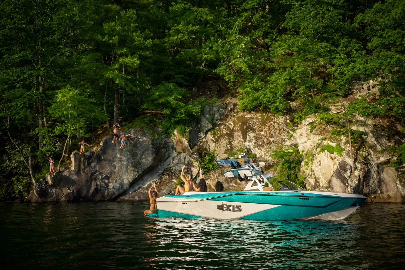 A teal Axis T250 idles beside a forested rock cliff as people leap from the rocks into the water, friends cheering aboard.